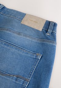 Jean en denim bleu clair avec un patch beige portant l'inscription "DENIM SUPPLY". Caractérisé par des coutures apparentes et une poche arrière. Texture lisse.