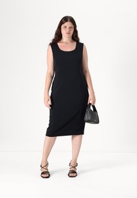 Abito midi nero senza maniche con scollo quadrato e vestibilità aderente. Abbinato a sandali neri con cinturini e una piccola borsa nera.