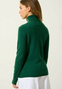 Mujer de espaldas hacia la izquierda, vistiendo un suéter de cuello alto verde oscuro y pantalones blancos, con cabello castaño de longitud media y pendientes de aro dorados.