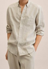 Chemise en lin gris clair avec col boutonné, poche poitrine unique et manches retroussées, présentant une coupe décontractée et une texture douce.