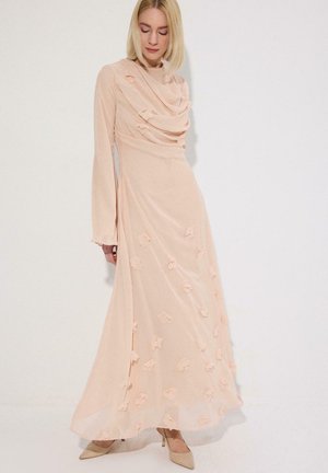 Γυναίκα που φοράει μακρύ, ρόζ-φούξια φόρεμα με υφασμάτινες λουλουδένιες λεπτομέρειες και μακριά μανίκια, συνδυασμένο με μπεζ μυτερά τακούνια, στέκεται μπροστά σε λευκό τοίχο.