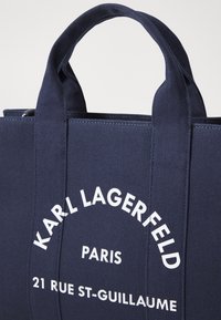 Marinblå canvas tote bag med dubbla handtag, med vit text: "KARL LAGERFELD PARIS 21 RUE ST-GUILLAUME." Stabil, strukturerad design.