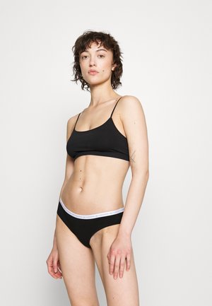 Čierny bavlnený bralet s tenkými ramienkami, kombinovaný s čiernymi bielizňovými nohavičkami v bikinovom štýle a bielym pásom s logom. Hladká textúra, minimalistický dizajn.