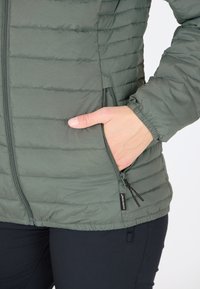 Vadderad jacka i salviagrön med horisontellt stygn. Har en ficka med dragkedja och elastiska mudd. Mjuk textur, lättviktig design.