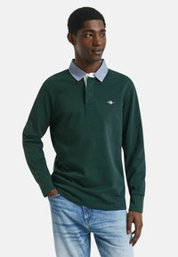 Junger Mann in einem dunkelgrünen Langarm-Polohemd mit hellblauem Kragen und hellblauen Jeans, der mit einer Hand in der Tasche steht.