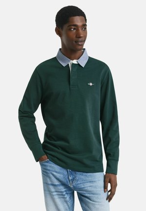 Young man wearing a dark green long-sleeve polo shirt with a light blue collar and light blue jeans, standing with one hand in pocket.