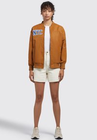 Giacca bomber marrone con ricamo "NYC" blu, con zip frontale, polsini a costine, indossata sopra un top bianco e pantaloni corti color crema, abbinata a scarpe da ginnastica.
