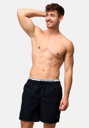 Shirtless young man smiling, wearing black drawstring shorts with visible white waistband, right hand resting on head against plain background.
