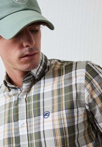 Gorra verde y camisa de cuadros con rayas verdes, marrones, azules y blancas, con botones y un emblema en el pecho. Material de algodón.
