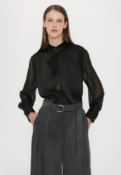 Black sheer blouse with a high collar and asymmetric placket, paired with gray tailored trousers featuring a wide waistband and belt.