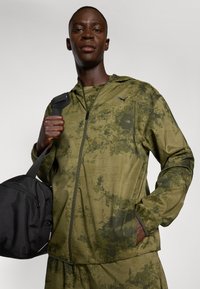Veste zippée vert olive avec un motif camouflé, manches longues et poches latérales. Le modèle tient un sac de sport noir.