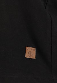 Schwarze Stoff mit gerippter Textur zeigt ein braunes quadratisches Lederpatch, das mit geprägtem Schriftzug "KEY LARGO" beschriftet ist.