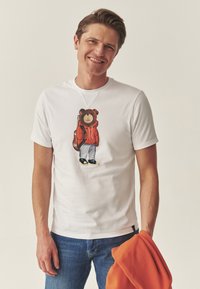 Fehér pamut póló, melyen egy medve grafikája látható, piros kapucnis pulóvert, kék farmert és sportcipőt viselve. A modell egy narancssárga pulóvert tart a kezében.