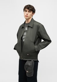 Νέος άνδρας που φοράει σκούρο πράσινο ζακέτα με φερμουάρ και μαύρο παντελόνι, με ένα ταίριαστο σκούρο καπέλο να κρέμεται από την τσέπη, στέκεται μπροστά σε ένα απλό φωτεινό φόντο.