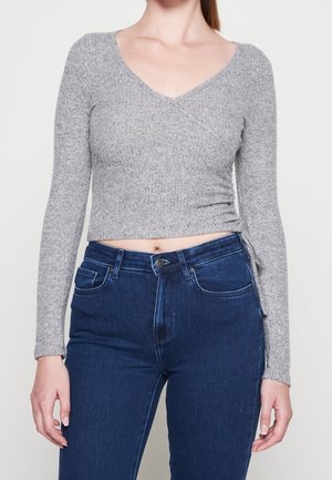 Frau trägt einen grauen, langärmeligen Wickelpullover und eine hoch taillierte dunkelblaue Jeans, steht vor einem einfarbigen hellen Hintergrund.