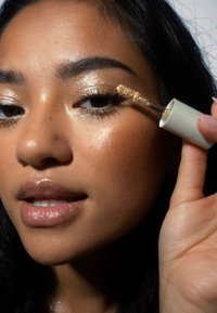 Primo piano di una persona che applica eyeliner glitter dorato con un tubetto bianco. La pelle ha una finitura lucida; le labbra sono di un beige lucido.