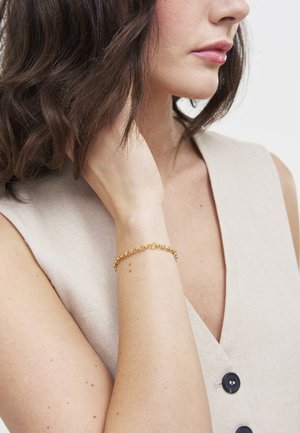 Žena s tamnom kosom duljine do ramena, odjevena u bež top bez rukava s crnim gumbima, dodirujući vrat, s narukvicom od zlatnog lanca na zapešću.