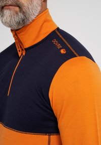 Orange og marineblå halv-zip pullover med høj krave. Har kontrasterende syninger og broderet brandlogo på skulderen. Blød tekstur.