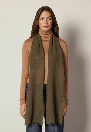 Femme portant un pull à col roulé marron, une écharpe vert olive drapée autour du cou et un jean bleu, debout devant un fond neutre.