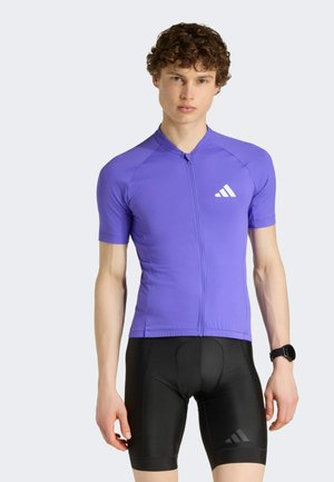 Junger Mann mit lockigem Haar, trägt ein enganliegendes lila Radsporttrikot und schwarze gepolsterte Radhosen, steht vor einem schlichten hellen Hintergrund.