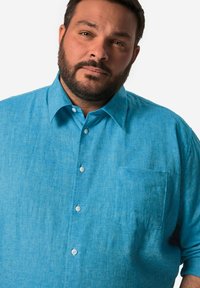 Světle modrá košile na knoflíky vyrobená z texturované tkaniny. Má límeček, nenápadnou kapsu a bílé knoflíky. Uvolněný střih s krátkými rukávy.