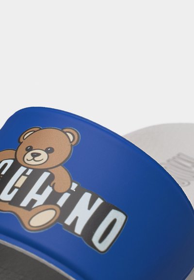 Μπλε διαφάνεια με γραφικό ενός χαμογελαστού αρκουδάκιου και μέρος του ονόματος της μάρκας «Moschino» με μεγάλα λευκά γράμματα.