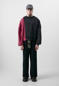 Übergroßes Sweatshirt in Schwarz und Burgunderrot mit abgewetzten Details, kombiniert mit schwarzen Hosen und klobigen schwarzen Sneakers. Graue Mütze mit Ösen.