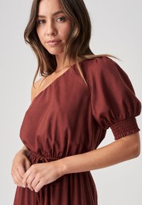 Sukienka typu off-shoulder w głębokim czerwonym kolorze. Posiada bufiaste rękawy ze smockowanym detalem oraz elastyczną talię z troczkami dla regulowanego dopasowania. Gładka tekstura materiału.