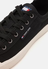 Svart lerret sneaker med runde lisser, sølvfargede øyer og en hvit gummisåle. Har en "TOMMY JEANS" -logo og et tri-farget detaljtag.