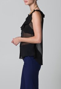 Top nero senza maniche con design arricciato, tessuto trasparente e una maglietta sottostante in tinta unita. Abbinato a pantaloni aderenti blu scuro. Accento a catena sulla spalla.