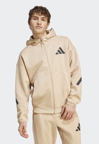 Sweatshirt bege com fecho éclair, feito de tecido texturizado, apresentando um grande logótipo preto no peito e detalhes pretos nas mangas.