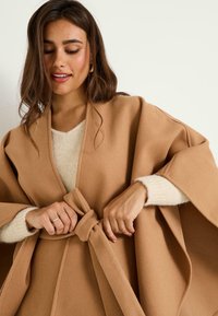 Kimono de estilo envolvente en tejido marrón y suave, con cintura ceñida con cinturón. Presenta mangas anchas y una cómoda blusa de textura beige debajo.