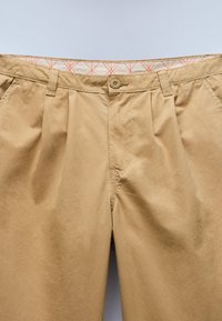 Khaki-colored casual pants with button closure, belt loops, side pockets, and a patterned inner waistband against a plain background.