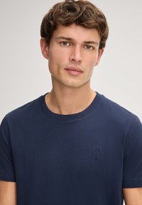 Marineblauwe katoenen t-shirt met korte mouwen en een ronde hals; heeft een subtiele bloemembroidery op de linkerkant van de borst. Glad textuur.
