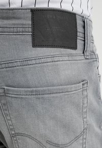 Primer plano del bolsillo trasero y la etiqueta de cuero negro en unos jeans Jack & Jones de color gris claro, usados con una camisa blanca a rayas.