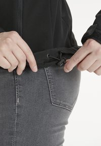 Svart jacka med drafsnodd i nederkant, som visar händer som justerar snodden. Bärs över grå denim, med textilstruktur och synliga sömmar.
