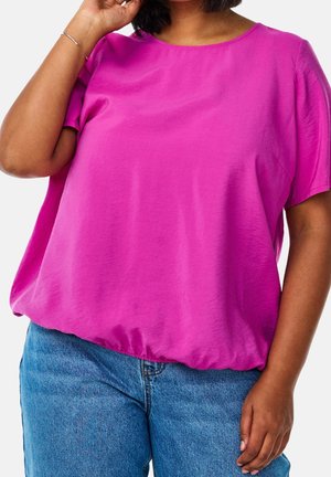 Nainen, joka pukeutunut löysään kirkkaan vaaleanpunaiseen lyhythihaiseen puseroon ja sinisiin farkkuihin, toinen käsi hiuksissa yksiväristä taustaa vasten.