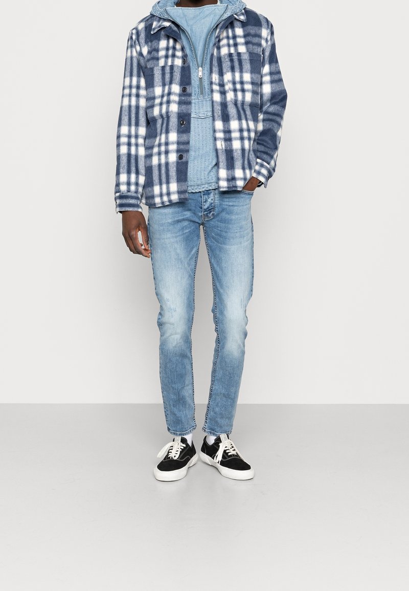 Personne portant un sweat à capuche bleu clair, une veste à carreaux bleue et blanche, un jean bleu délavé et des baskets noires et blanches debout sur un sol gris.