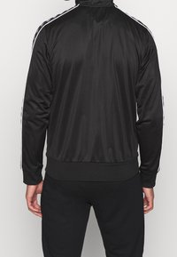 Chaqueta de chándal negra hecha de material suave, con rayas blancas a lo largo de los hombros y una cinturilla acanalada para un ajuste ceñido.