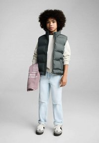 Gilet imbottito grigio sopra un maglione crema, jeans azzurri con strappi e sneakers grigie. Tiene una felpa rosa con testo bianco.