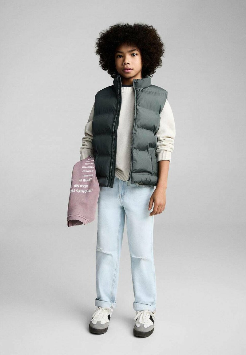 Gilet imbottito grigio sopra un maglione crema, jeans azzurri con strappi e sneakers grigie. Tiene una felpa rosa con testo bianco.