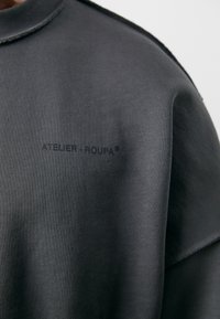 Sweatshirt oversize en gris charbon fabriqué dans un tissu doux, avec une texture côtelée subtile et le logo "ATELIER-ROUPA" en impression noire.