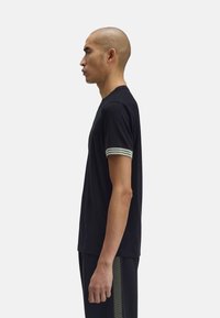 T-shirt nera a maniche corte con collo tondo, caratterizzata da accenti a strisce verdi e bianche sui polsini. Il tessuto appare morbido e liscio.