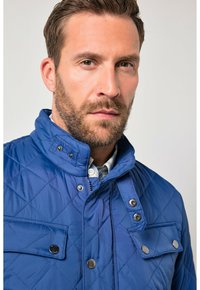 Niebieska pikowana kurtka z wysokim kołnierzem, zatrzaskami i kieszenią na piersi z guzikiem. Gładka, lekka tkanina.