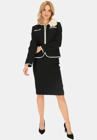 Czarny garnitur z tkaniny tweedowej z białymi detalami, dopasowana marynarka z dwiema kieszeniami i akcentem w postaci kwiatu, zestawiona z ołówkową spódnicą i czarnymi szpilkami.
