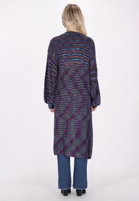 Lång, oversized cardigan i lila och blå nyanser med randigt mönster, stickad yttextur och ballongsleeves. Bärs över flarejeans.