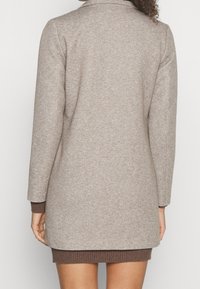 Dlouhý béžový pletený kabát s jemným texturovaným vzorem. Má šálový límec a hnědé žebrované detaily na manžetách a lemu.