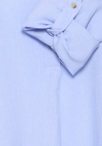 Poignet de chemise à manches longues bleu clair retroussé et boutonné, montrant un tissu tissé texturé et un détail de bouton beige.