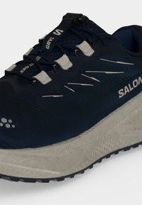 Σκούρο μπλε παπούτσι trail της Salomon με λευκή σόλα και σύστημα γρήγορου κορδονιού σε λευκό φόντο.