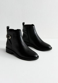 Botas de tobillo negras de cuero suave con punta afilada, cierre lateral de cremallera y detalle de hebilla dorada en el lado exterior. Tacón bajo tipo bloque.